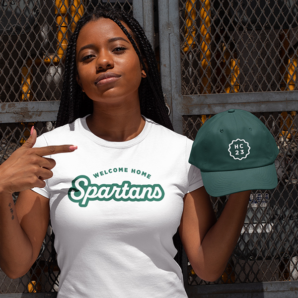 Woman wearing t-shirt reading "Welcome Home Spartans" and holding MSU Homecoming 2023 branded hat.