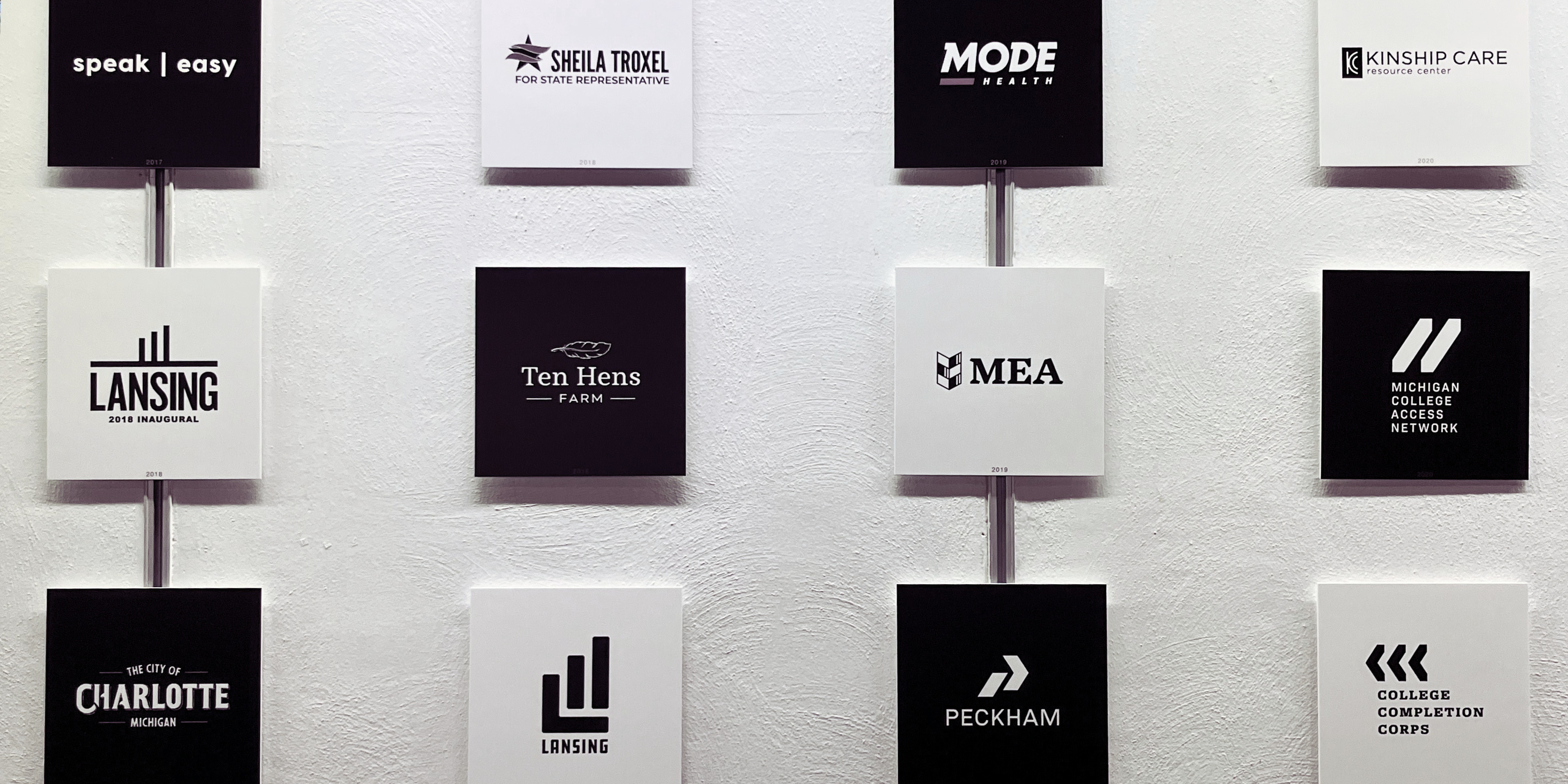 Logos created by Redhead are displayed on a wall with alternating black and white backgrounds. Some include Ten Hens Farm, City of Lansing, and MEA